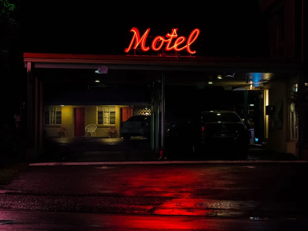 A red neon "Motel" sign glows above a dark parking area and guest rooms at night